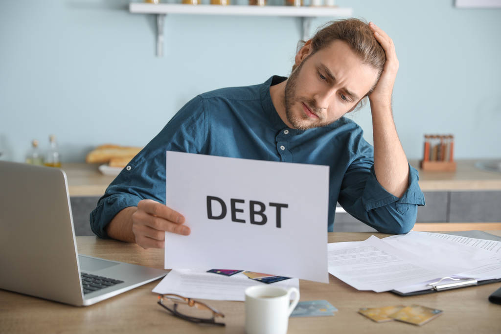 A stressed man at a kitchen table holding a paper marked debt while surrounded by bills and credit cards